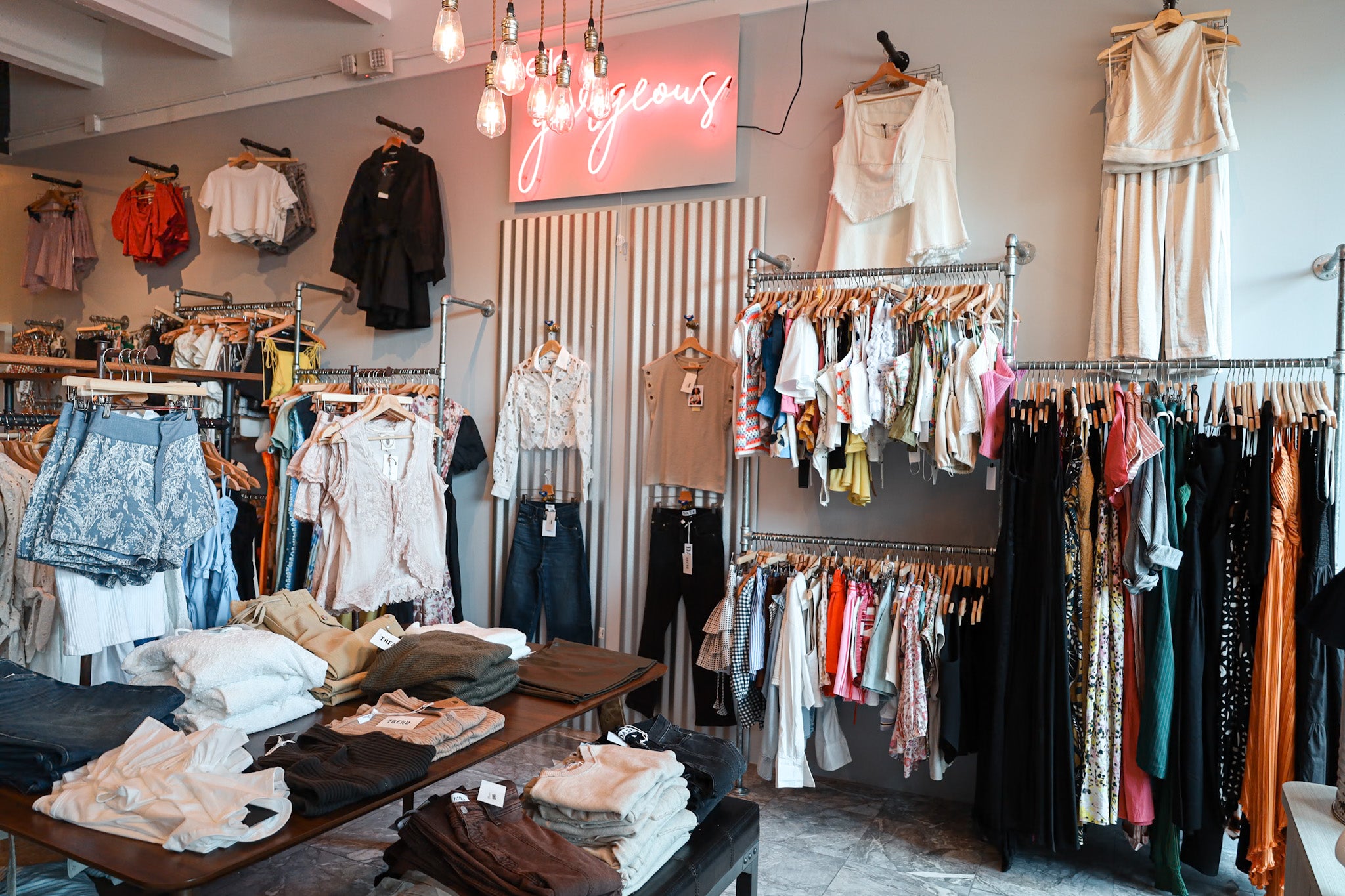 Inside an IF SO boutique: racks of tops and dresses under a pink neon sign that reads gorgeous, with folded denim on a center table